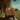 Two children sit on top of hay bales loaded onto a truck driving down a paved road, viewed from behind. The children are facing forward with their backs to the camera, the hay bales are golden brown, and the background features green fields and trees under a blue sky with some wispy clouds. The image evokes a sense of nostalgia and the joy of childhood.