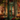 A charming shop storefront is illuminated at night with warm Christmas lights. The dark green building is decorated with garlands of evergreen, red berries, and twinkling fairy lights draped across the windows and above the ornate double doors, which also bear a red berry wreath. Large display windows reveal brightly lit interiors filled with more holiday decor, while potted red berry plants and wrapped presents sit on the sidewalk, creating a festive scene.