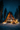 A stunning low-angle shot of a warmly lit A-frame cabin situated in a deep snow-covered forest at night. The cabin's entire front facade is made of glass, glowing with golden light, and is flanked by tall, dark pine trees dusted with snow. Festive Christmas trees adorned with bright golden lights stand prominently in the snow on either side of the cabin's entrance, beneath a clear, star-filled dark blue night sky.