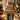 A person is shown from the chest down, wearing a cream-colored cable-knit sweater with a textured pattern and light blue jeans. They are holding a vertical stack of four wrapped presents, three in plain brown kraft paper with natural twine, and one in white paper with gold speckles and twine. The background is softly blurred with warm, golden bokeh lights from a Christmas tree and other holiday decorations, creating a festive and cozy indoor scene.