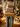 A person is shown from the chest down, wearing a cream-colored cable-knit sweater with a textured pattern and light blue jeans. They are holding a vertical stack of four wrapped presents, three in plain brown kraft paper with natural twine, and one in white paper with gold speckles and twine. The background is softly blurred with warm, golden bokeh lights from a Christmas tree and other holiday decorations, creating a festive and cozy indoor scene.