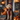 A medium-sized golden doodle dog sits on stone steps, facing forward and dressed in a vampire costume consisting of a red vest and a black cape with a high collar. The dog's golden fur contrasts with the dark costume. The house steps are decorated with multiple orange pumpkins and metal framed lanterns with flickering candles, creating a Halloween scene.