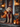 A medium-sized golden doodle dog sits on stone steps, facing forward and dressed in a vampire costume consisting of a red vest and a black cape with a high collar. The dog's golden fur contrasts with the dark costume. The house steps are decorated with multiple orange pumpkins and metal framed lanterns with flickering candles, creating a Halloween scene.