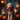 A medium-sized Goldendoodle dog sits attentively in a portrait style photograph wearing a vampire costume. The dog is dressed in a black cape with a red lining, a red vest, and a bowtie. The dog is sitting on a patterned rug in front of wooden porch and lanterns with pumpkins on either side.