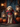 A medium-sized Goldendoodle dog sits attentively in a portrait style photograph wearing a vampire costume. The dog is dressed in a black cape with a red lining, a red vest, and a bowtie. The dog is sitting on a patterned rug in front of wooden porch and lanterns with pumpkins on either side.