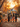 Five young girls in Halloween costumes walk away from the viewer down a residential street covered in autumn leaves. The girls, dressed as various kinds of witches, carry pumpkin-shaped candy buckets. The scene is bathed in warm, golden light from the setting sun, and decorated with lit streetlights and jack-o-lanterns on the curbside.