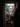 Five children in Halloween costumes stand at the threshold of a doorway, eagerly awaiting candy. Three of the children are in the front, holding trick-or-treat bags, while two others peek out from behind. A blurred figure stands in the foreground on the right, presumably the homeowner about to open the door. Outside, a suburban neighborhood is visible with a white house and trees with autumn foliage.
