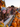 A group of six teenage friends are smiling at the camera, posing behind a wooden fence at a pumpkin patch. The fence is lined with hay bales and a multitude of pumpkins in various sizes fill the foreground. Bright orange and yellow fall flowers decorate the background and warm string lights add to the festive atmosphere.