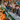 Six teenagers, of various ethnicities, are smiling while leaning on a gray wooden fence. They are surrounded by many orange pumpkins and colorful autumn flowers, with a red barn visible blurred in the background. The image evokes a feeling of fall and friendship at an outdoor festival.