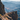 A wide shot of a group of five hikers walking single-file along a rocky mountain ridge. The hikers are seen from behind and are silhouetted against a backdrop of layered mountains in varying shades of blue and brown. The sky is clear and blue, and the overall scene conveys a sense of vastness and adventure.