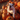A close-up photograph shows a white and brown bulldog puppy with a happy expression, sitting on top of several bright orange pumpkins. The pumpkins are of varying sizes and shapes, forming a pile on hay. The puppy is facing the camera with its tongue slightly out, and more pumpkins fill the blurred background.