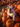 A close-up photograph shows a white and brown bulldog puppy with a happy expression, sitting on top of several bright orange pumpkins. The pumpkins are of varying sizes and shapes, forming a pile on hay. The puppy is facing the camera with its tongue slightly out, and more pumpkins fill the blurred background.