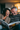 A middle-aged woman and man are sitting closely together, engrossed in a magazine. The woman, wearing a knitted sweater, is laughing heartily with her eyes closed. The man, with graying hair, is smiling broadly, his eyes sparkling with amusement as he looks at the magazine. The warm, indoor setting creates a sense of intimacy and joy.