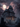 A large, three-story gothic mansion sits amidst a graveyard under a full moon. The house is gray and imposing, with many windows and a covered porch. Gravestones of various shapes and sizes dot the foreground, and a flock of birds flies across the stormy, pink-tinged sky.