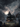 A digitally rendered image shows a three-story gothic mansion on a grassy lot, under a dramatic sky filled with dark clouds and a bright full moon. Gravestones are scattered in front of the house. Silhouetted birds fly through the sky, adding to the ominous and eerie atmosphere.