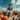 A scarecrow wearing denim overalls, a plaid shirt, and a straw hat sits on top of a hay bale inside a wooden wagon. The wagon also contains numerous pumpkins and a pot of vibrant orange flowers. The background features a clear blue sky dotted with fluffy white clouds, suggesting a bright, sunny autumn day.