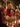 A blonde little girl in a red knitted sweater sits on a man's shoulders, both laughing joyfully at a festive, candlelit dinner table. The man, with dark hair and a patterned beige sweater, smiles broadly, surrounded by other family members blurred in the foreground and background. Warm, glowing Christmas lights create a cozy and celebratory atmosphere, highlighting the genuine connection and happiness shared during the holiday meal.