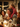A father with curly hair and a beard, wearing a patterned knit sweater, smiles broadly while carrying his young daughter on his shoulders. The daughter, also smiling brightly and wearing a red knit sweater, looks to her right with genuine happiness. They are seated at a dimly lit festive dinner table adorned with glowing candles and blurred string lights, with another smiling woman in a festive sweater visible in the background.
