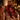 A young girl with a bright smile sits on a man's shoulders, both laughing, at a festive dinner table. A woman with blonde hair smiles on the left, with warm candle and blurred Christmas tree lights illuminating the cozy, evening scene. All three individuals are wearing cozy winter sweaters.