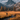A person with a backpack walks through an autumn meadow with a stream running through it. Behind the person is a large mountain range with gray, jagged peaks under a partly cloudy sky. The meadow is covered in golden grasses and some autumn-colored trees, with the overall impression of a serene and adventurous outdoor scene.