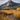 A scenic landscape image shows a lone hiker walking away from the viewer across a golden meadow toward a large mountain in the distance. A small stream flows in the foreground, with its clear water reflecting the sky. The landscape is painted with autumn colors of yellow, gold, and green, under a blue sky dotted with white clouds, creating a sense of vastness and peacefulness.