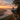 A hiker with a backpack stands on the edge of a rocky cliff, looking out at a mountain range shrouded in fog during sunrise. The scene is bathed in warm, golden light, with the sky filled with soft clouds. Pine trees frame the right side of the image.