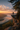 A hiker with a backpack stands on the edge of a rocky cliff, looking out at a mountain range shrouded in fog during sunrise. The scene is bathed in warm, golden light, with the sky filled with soft clouds. Pine trees frame the right side of the image.