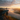 A hiker with a backpack stands on the edge of a rocky cliff, looking out over a mountain valley filled with mist. The scene is illuminated by a soft, warm light as the sun rises or sets, creating a breathtaking view of layered mountains, a river, and a partially cloudy sky.