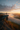 A hiker with a backpack stands on the edge of a rocky cliff, looking out over a mountain valley filled with mist. The scene is illuminated by a soft, warm light as the sun rises or sets, creating a breathtaking view of layered mountains, a river, and a partially cloudy sky.