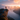 A person with a backpack stands on the edge of a rocky cliff. Below them is a large lake surrounded by forested mountains, partially obscured by mist. The sky above is colored with the hues of a rising sun, with pink and orange clouds highlighting the scene.