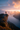 A person with a backpack stands on the edge of a rocky cliff. Below them is a large lake surrounded by forested mountains, partially obscured by mist. The sky above is colored with the hues of a rising sun, with pink and orange clouds highlighting the scene.