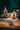 A charming light-colored brick house at night during winter, covered in snow and adorned with festive Christmas decorations. A large evergreen tree on the left is illuminated with multi-colored lights, while the front door of the house is decorated with green garlands, red poinsettias, and outlined with warm white lights, creating a cozy and inviting scene.