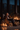 A silver haired man wearing glasses sits in a gray armchair reading a book and holding a glass of red wine. A golden retriever lies at his feet on a rug. Behind him, a modern fireplace with burning logs glows warmly. To the left, various glass candle holders filled with lit candles add to the warm glow of the scene.