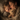A mother and child are sleeping in bed with a teddy bear. The mother has dark hair and fair skin, and her eyes are closed. The child, who appears to be a toddler, has blond hair and is also sleeping soundly with their eyes closed. A tan-colored teddy bear rests in their arms. They are snuggled beneath a light-colored blanket in a dimly lit room.