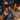 A mother and her two young daughters are sitting close together on a gray couch, covered with a beige blanket. They are all smiling and looking to the right of the frame, presumably watching television. Soft candlelight illuminates the room, creating a warm and inviting atmosphere.