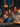 A mother and her two young daughters are sitting close together on a gray couch, covered with a beige blanket. They are all smiling and looking to the right of the frame, presumably watching television. Soft candlelight illuminates the room, creating a warm and inviting atmosphere.