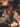 A smiling mother sits on a couch with her two young daughters. They are snuggled together, partially covered by a cream-colored blanket. The scene is softly lit by the warm glow of numerous candles, creating a cozy and inviting atmosphere.