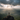 A person stands on top of a rocky mountain peak with arms raised in celebration. The sun shines through the clouds in the background, casting rays of light over a green valley filled with trees. The person is silhouetted against the bright sky, conveying a sense of triumph and connection with nature.