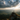 A person stands silhouetted with arms raised on the peak of a rocky mountain at sunset. The person faces the setting sun, whose golden rays stream through a partly cloudy sky above a vast landscape of rolling green hills and a dark green forest. The scene conveys a sense of triumph and connection with nature.