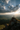 A person stands silhouetted with arms raised on the peak of a rocky mountain at sunset. The person faces the setting sun, whose golden rays stream through a partly cloudy sky above a vast landscape of rolling green hills and a dark green forest. The scene conveys a sense of triumph and connection with nature.