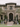 A wide shot of a grand, two-story house decorated for Halloween. Skeletons, pumpkins, and tombstones line the front steps and lawn. Three people dressed in dark gowns stand on the steps, and a person in white stands on the balcony, overlooking the scene. The house is tan with dark gray accents and surrounded by palm trees.