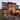 A large house is decorated for Halloween with multiple tombstones, cacti, and Halloween figures in the front yard. Several people in costume stand on the steps leading up to the arched entrance. The house is lit with warm light, creating an inviting atmosphere against the dusky sky.
