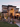 A large house is decorated for Halloween with multiple tombstones, cacti, and Halloween figures in the front yard. Several people in costume stand on the steps leading up to the arched entrance. The house is lit with warm light, creating an inviting atmosphere against the dusky sky.