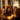 A group of people are seated around a dining table during a Thanksgiving dinner. The warm light from candles illuminates the table and the faces of people engaging in conversation. An arrangement of Autumnal colored flowers is at the center. The setting appears to be a home interior decorated for the season.