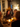 A group of people are seated around a dining table during a Thanksgiving dinner. The warm light from candles illuminates the table and the faces of people engaging in conversation. An arrangement of Autumnal colored flowers is at the center. The setting appears to be a home interior decorated for the season.