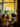 A group of four young women are seated around a wooden table, engaging in conversation during an autumn celebration. One woman, positioned near the window, smiles warmly, illuminated by soft natural light. Through the window, vibrant autumn foliage with orange and yellow leaves can be seen.