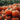 Close-up shot of a large group of bright orange pumpkins with green stems, piled on top of each other in a field. In the background, a blurred expanse of green foliage creates a natural backdrop. The overall scene suggests a pumpkin patch during the fall harvest season.
