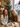 A medium shot captures two young women, one with red hair and another with blonde curly hair, laughing heartily at a wooden table in a festive cafe. In front of them are a latte, a slice of cake, and glowing candles, with a decorated Christmas tree and string lights in the warm, blurred background, and a street scene visible through a window. The image conveys genuine joy and a cozy holiday atmosphere.