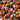 A close-up photograph shows a dense, colorful mix of Halloween candy. The candies include classic candy corn with yellow, orange, and white stripes, orange pumpkin-shaped candies with carved black faces, and round gumball-like candies decorated with various Halloween-themed designs such as eyeballs, ghosts, bats, and monster faces. The candies are tightly packed, filling the entire frame.