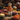 A scarecrow stands in the center of the frame, surrounded by a vast array of colorful potted mums. The scarecrow has a straw head with a hand-drawn smiling face, an orange plaid shirt, and white pants with straw accents. Several pumpkins sit at the base of the scarecrow on a bed of hay, contributing to the overall autumn harvest theme.